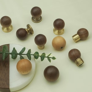 Pode incluir: Uma coleção de puxadores de armário de madeira em vários estilos. Alguns puxadores são castanhos escuros com bases de latão, enquanto outros são de madeira clara. Os puxadores estão dispostos sobre uma superfície verde clara com uma planta decorativa e um porta-copos de mármore e madeira.