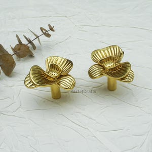 Könnte beinhalten: Zwei goldfarbene, blütenförmige Schrankknöpfe. Jeder Knopf hat einen Stiel und ein detailliertes Blumendesign. Die Knöpfe befinden sich auf einer weißen Oberfläche mit einem Zweig aus getrockneten Blättern im Hintergrund. Der Text "PerfetcCrafts" ist sichtbar.