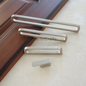 Op de afbeelding: Een verzameling zilverkleurige kastbeslagen, waaronder lange stangtrekkers en een kleine handgreep. De hardware wordt getoond tegen een donkere houten deur en een lichtgekleurd oppervlak. De tekst "PerfecCrafts" is zichtbaar.