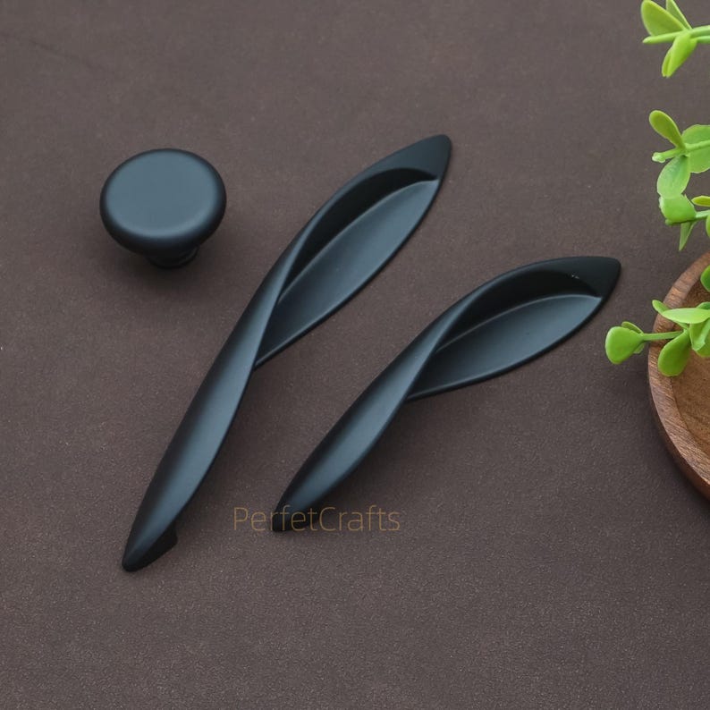 以下が含まれることがあります： 丸いノブと2つの湾曲したハンドルを備えた黒いキャビネットハードウェアセット。 ハードウェアはマット仕上げでモダンなデザインです。 ブランド名「PerfetcCrafts」が右下に表示されています。