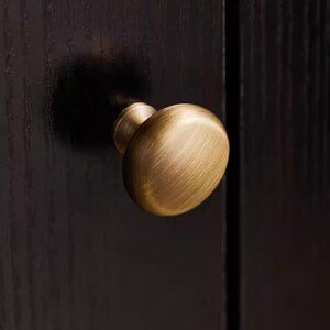K&ouml;nnte beinhalten: Nahaufnahme eines Messing-Schrankknaufs mit ovaler Form. Der Knauf ist an einer dunkelbraunen Holzschrankt&uuml;r befestigt. Der Knauf hat eine warme, antike Oberfl&auml;che, und die Holzmaserung ist sichtbar.