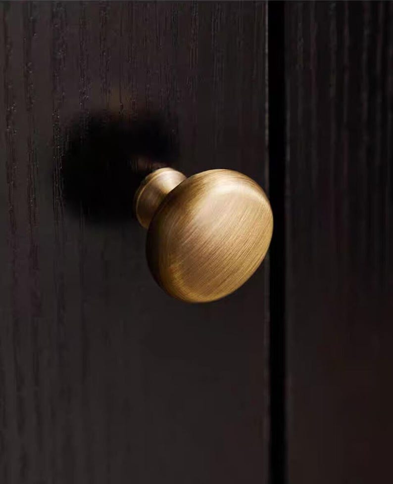 K&ouml;nnte beinhalten: Nahaufnahme eines Messing-Schrankknaufs mit ovaler Form. Der Knauf ist an einer dunkelbraunen Holzschrankt&uuml;r befestigt. Der Knauf hat eine warme, antike Oberfl&auml;che, und die Holzmaserung ist sichtbar.