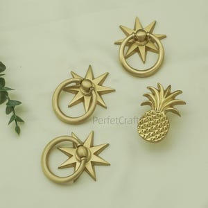 May include: Four gold-coloured cabinet pulls. Three pulls feature a starburst design with a ring handle, and one is shaped like a pineapple. The text "PerfektCraft" is visible on the surface.