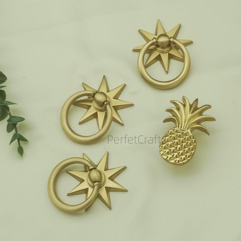 May include: Four gold-coloured cabinet pulls. Three pulls feature a starburst design with a ring handle, and one is shaped like a pineapple. The text "PerfektCraft" is visible on the surface.