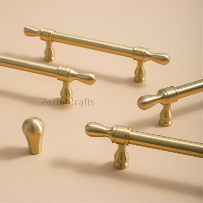 May include: A selection of gold-coloured cabinet hardware, including several bar pulls and a single knob. The pulls have a simple, elegant design with a cylindrical bar and decorative ends. The word "PerfectCrafts" is printed on the surface.