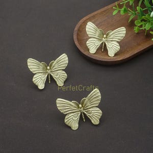 K&ouml;nnte beinhalten: Drei goldfarbene, schmetterlingsf&ouml;rmige Dekorationsgegenst&auml;nde. Die Schmetterlinge haben detaillierte Fl&uuml;gelmuster und sind auf einer dunkelbraunen Oberfl&auml;che angeordnet. Ein Schmetterling befindet sich auf einem Holztablett. Der Text "PerfecCrafts" ist sichtbar.