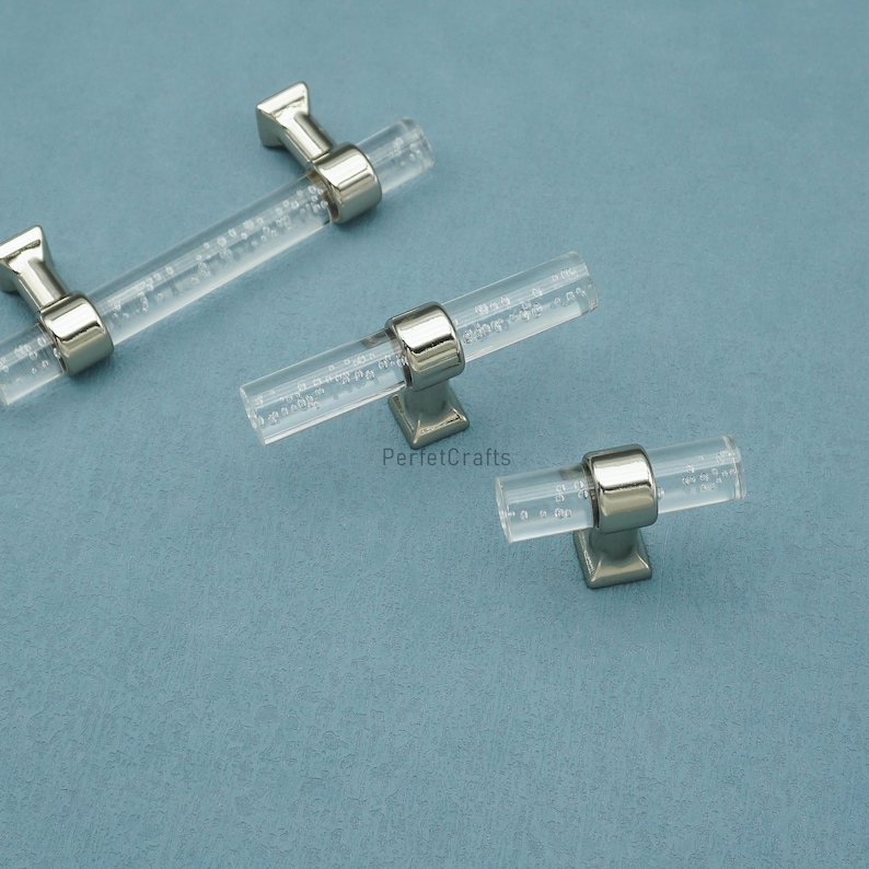 Peut inclure: Poign&eacute;es de meubles en acrylique transparent avec des accents m&eacute;talliques argent&eacute;s. Les poign&eacute;es ont un design cylindrique avec de petites bulles &agrave; l'int&eacute;rieur de l'acrylique. Le mat&eacute;riel est pr&eacute;sent&eacute; sur une surface bleu clair. Le texte "PerfetcCrafts" est visible.