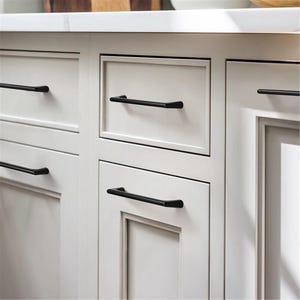 May include: Close-up of white kitchen cabinets with black bar handles. The cabinets have a recessed panel design and are arranged in a row. The black handles are horizontally mounted on the drawers and doors.