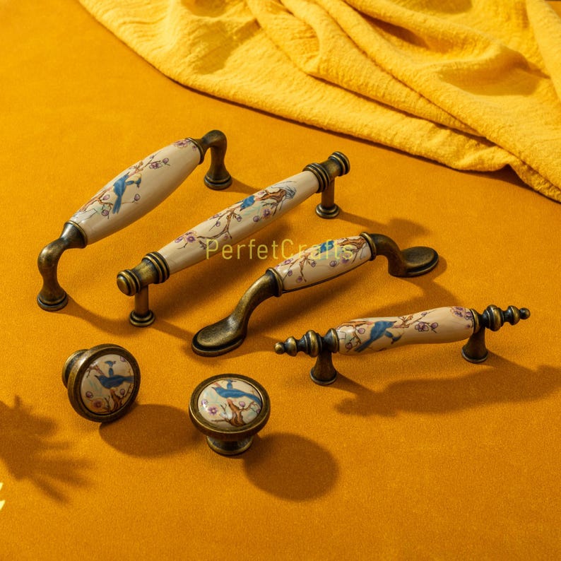 May include: A collection of ceramic cabinet pulls and knobs. The pulls have a cream-colored base with a blue bird and floral design, and bronze-colored metal hardware. The knobs have a matching bird and floral design. The word "PerfetcCrafts" is printed on the pulls.