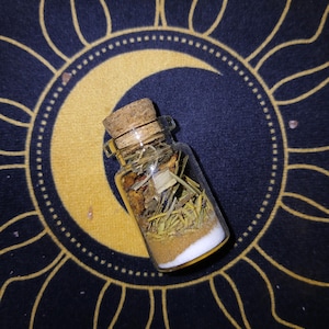 Puede incluir: Una pequeña botella de vidrio transparente llena de capas de hierbas y polvos, sellada con un tapón de corcho. La botella descansa sobre una superficie azul oscuro con un diseño dorado de sol y luna.