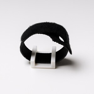 May include: A black hook and loop cable tie, secured by a white plastic holder. The tie is made of a soft, textured material and is looped around itself. The background is plain white.