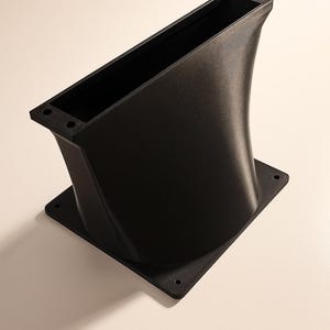 Puede incluir: Un objeto negro, angular y rectangular con un lado curvado y ensanchado. El objeto tiene una base cuadrada y una abertura rectangular en la parte superior. El objeto parece estar hecho de un material negro mate.