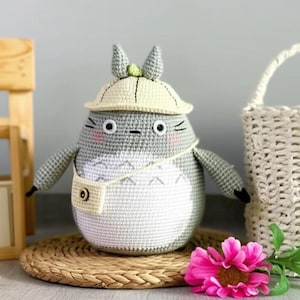 Puede incluir: Un peluche de Totoro hecho a mano con ganchillo, con cuerpo gris, vientre blanco y sombrero amarillo. El peluche tiene brazos pequeños, ojos negros y mejillas rosadas. Lleva una pequeña bolsa blanca. Una flor rosa y una estera tejida están en primer plano.