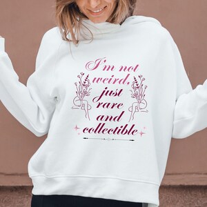 Puede incluir: Sudadera con capucha blanca con el texto "I'm not weird, just rare and collectible" en fuente cursiva, con ilustraciones florales. La sudadera con capucha la lleva una persona. El fondo es de color marrón liso.