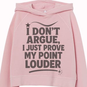 Peut inclure: Un sweat à capuche rose clair avec le texte "I DON'T ARGUE, I JUST PROVE MY POINT LOUDER" en gris foncé. Le sweat-shirt comporte une étoile et un motif de soleil. La marque "Oneder Wall Art" est visible.