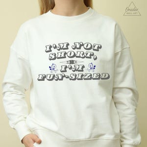 Pode incluir: Camisola branca com o texto "I'M NOT SHORT, I'M FUN-SIZED" em cinza, contornado a preto, com elementos decorativos azuis. A camisola tem gola redonda e mangas compridas. A marca "ONEDER WALL ART" está no canto superior direito.