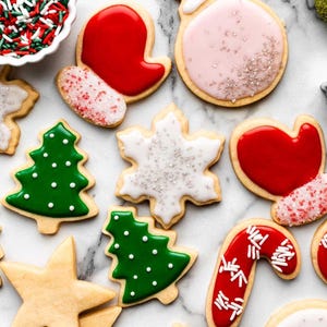 Puede incluir: Vista aérea de galletas navideñas variadas con glaseado colorido. Las formas incluyen un árbol de Navidad verde, un guante rojo, un copo de nieve, una bola de Navidad, un corazón y un bastón de caramelo. Se ven chispas en un cuenco blanco pequeño.