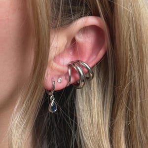 May include: An ear featuring multiple silver earrings. Three stacked hoop earrings are on the upper ear, a small stud, and a dangling earring with a teardrop-shaped, dark blue stone.