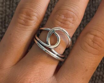 May include: A silver ring with a unique, intertwined design. The ring features a central loop and multiple bands, creating an abstract, modern look. The hand wearing the ring has light pink nail polish.