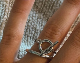 May include: A silver ring with a toggle clasp design. The ring features a circular element connected to a straight bar, creating a modern and minimalist aesthetic. The ring is worn on a finger.