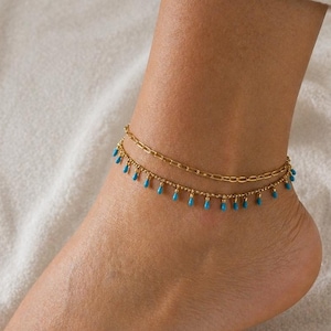 May include: A pair of gold anklets are worn on an ankle. The upper anklet is a plain chain, and the lower one has small, turquoise teardrop beads. These anklets offer a touch of elegance and are a stylish accessory.