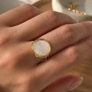 May include: A gold ring featuring a circular, white mother-of-pearl stone. The ring is worn on a hand, with a white sleeve visible. The ring has a textured gold band around the stone, offering a distinctive and refined aesthetic.