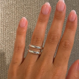 May include: A hand with oval-shaped nails featuring a soft pink and white ombre design. A double-banded silver ring adorns the ring finger. The nails have a glossy finish, and the skin tone is light.