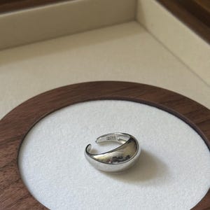 May include: A silver ring with a smooth, curved design. The ring has an open, adjustable band. The ring is displayed on a white surface within a wooden frame.