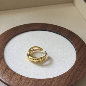 Puede incluir: Un anillo dorado con un diseño en capas. El anillo presenta un acabado liso y pulido y una forma curva. El anillo se muestra sobre una superficie blanca dentro de un marco de madera.