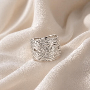 May include: A silver ring with a woven design, set with small, sparkling stones. The ring is displayed on a cream-coloured, silky fabric, creating a luxurious and elegant aesthetic. The ring's intricate pattern and bright finish make it a striking piece of jewellery.