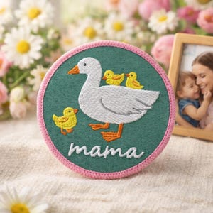 May include: Embroidered patch featuring a white goose with three yellow ducklings on a teal background, framed by a pink border. The word "mama" is stitched in white. The patch is round and appears to be made of fabric.