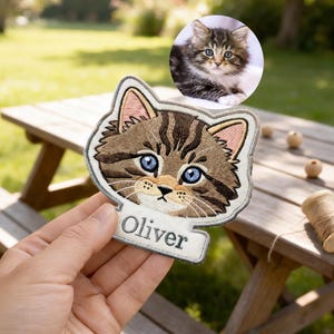 May include: Embroidered patch featuring a tabby cat face with blue eyes and the name "Oliver" on a banner. The patch is held in a hand, with a small photo of a similar cat in the background.