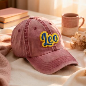 May include: A dusty rose baseball cap with the word "Leo" embroidered in yellow and blue. The cap has a curved brim and a distressed look. A mug and a stack of books are in the background.