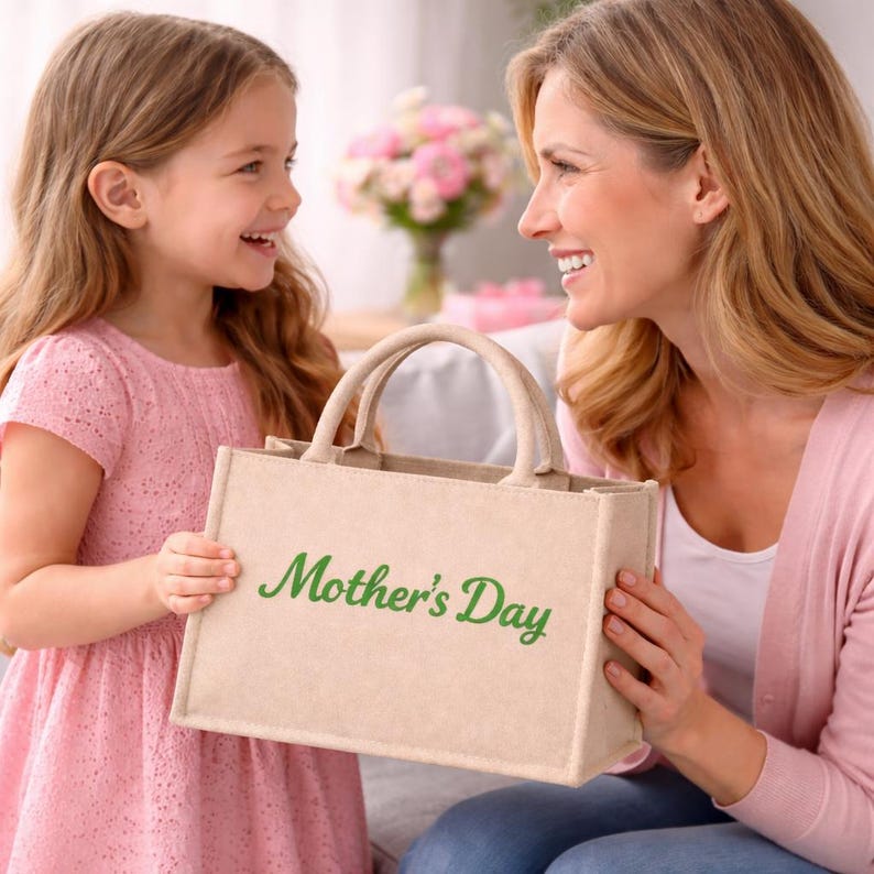 May include: A beige tote bag with the words "Mother's Day" in green script. The bag has two handles and is being held by a woman and a young girl. The girl is wearing a pink dress.
