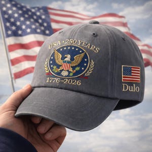 May include: Gray baseball cap with an embroidered emblem featuring a bald eagle, stars, and the text "USA 250 YEARS 1776-2026." A small American flag patch and the name "Dulo" are on the side. The U.S. flag is in the background.