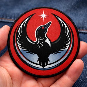 May include: Round patch featuring a stylized black bird with outstretched wings, set against a red background with a white star. The bird has a red eye and white accents. The patch has a red border and a blue and white gradient at the bottom.