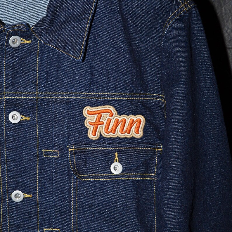 May include: A dark blue denim jacket with a collared neckline, button closures, and a chest pocket. The jacket features the name "Finn" embroidered in orange and tan on the chest.