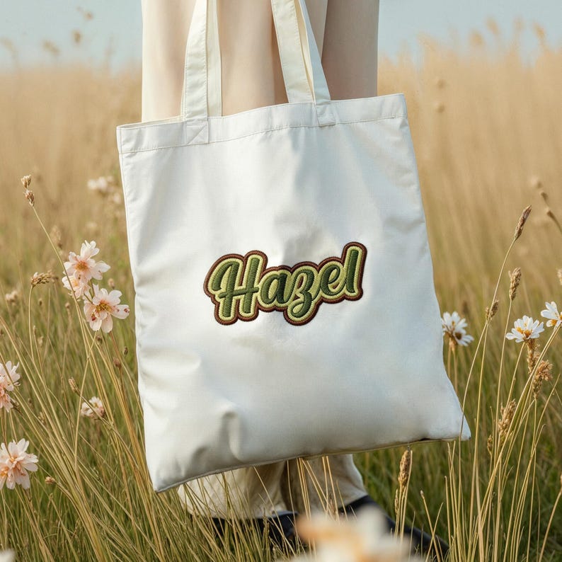 May include: A white tote bag with the name "Hazel" embroidered in green and brown script. The bag is held in a field of tall grass and wildflowers, suggesting an outdoor or nature-themed product.