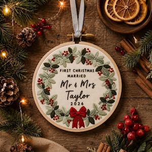 May include: A round wooden embroidery hoop containing a cream-colored fabric with a hand-stitched wreath of green leaves and red berries. The text reads FIRST CHRISTMAS MARRIED Mr & Mrs Taylor 2026, with a red fabric bow at the bottom.