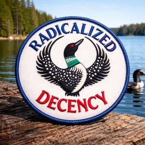 May include: Round patch with a white background, blue border, and the words "RADICALIZED DECENCY" in blue and red. The patch features a stylized black and white bird with a green and black neck. A lake and trees are in the background.