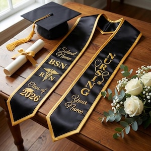 May include: A graduation cap with a gold tassel, diploma, and two black graduation stoles with gold trim. The stoles are embroidered with "School Name BSN R N Class of 2026" and "NURSING Your Name". A bouquet of white roses is also present.