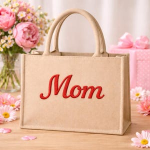 May include: A beige tote bag with the red embroidered word "Mom" on the front. The bag has two handles. Pink flowers and a gift box are in the background. The bag is on a wooden surface.