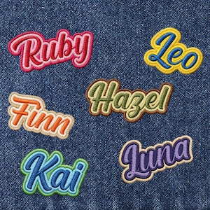 May include: Embroidered name patches in various colors, including Ruby, Leo, Finn, Hazel, Kai, and Luna. Each patch features a unique color scheme and a cursive font, perfect for personalizing clothing or accessories. The patches are set against a denim background.