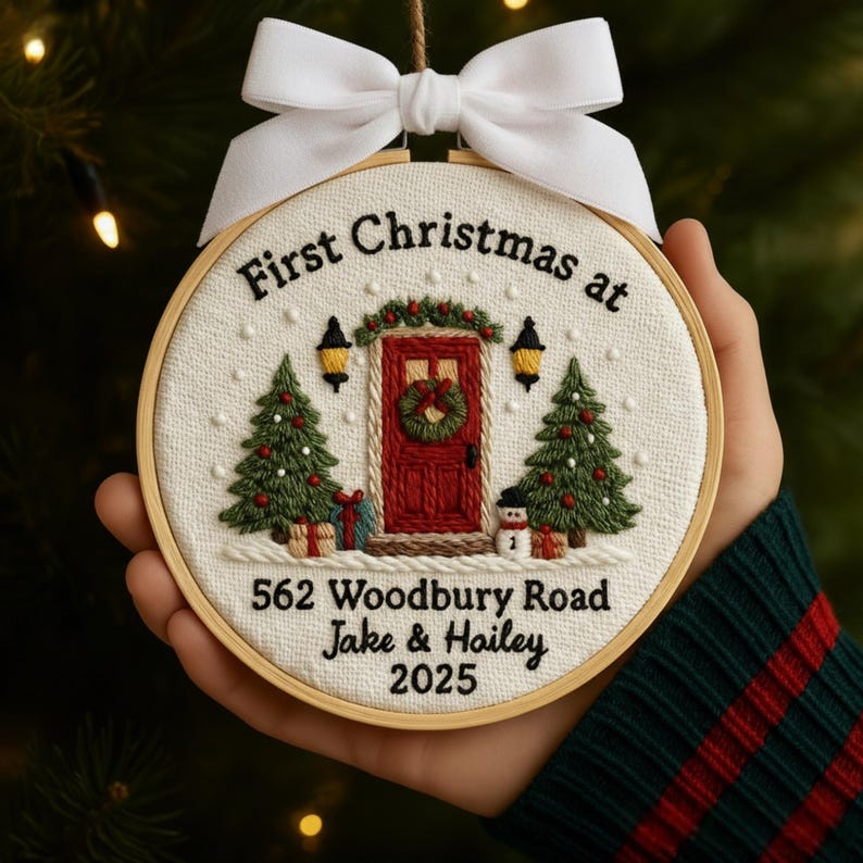 May include: Embroidered Christmas ornament in a wooden hoop. The design features a red door with a wreath, Christmas trees, and the text "First Christmas at 562 Woodbury Road Jake & Hailey 2025". A white bow is at the top.