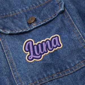 May include: Close-up of a denim jacket pocket with a purple and tan embroidered "Luna" patch. The jacket is a medium wash blue with visible stitching and a metal button.