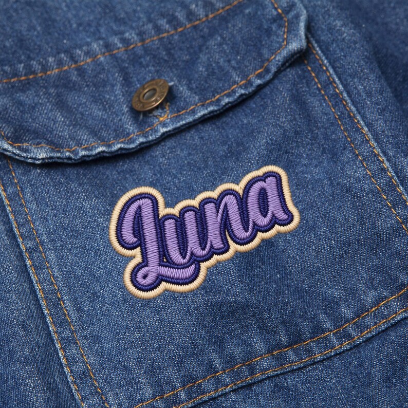 May include: Close-up of a denim jacket pocket with a purple and tan embroidered "Luna" patch. The jacket is a medium wash blue with visible stitching and a metal button.
