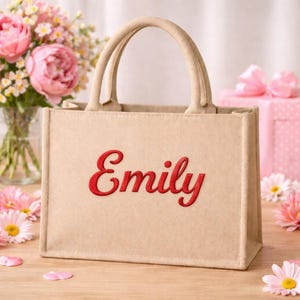 May include: A beige tote bag with two handles and the name "Emily" embroidered in red script. The bag is rectangular and sits on a wooden surface, with flowers and a gift box in the background.
