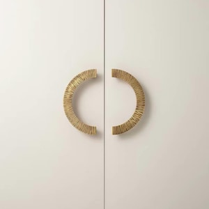 May include: Two semi-circular cabinet pulls in a metallic gold finish, each featuring a textured, ribbed surface. The handles are mounted symmetrically on a pair of flat, light-colored cabinet doors, creating a circular shape when closed together.
