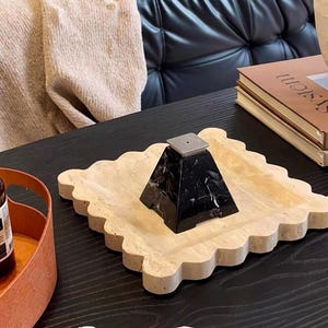 May include: A decorative tray with a scalloped edge holds a black marble pyramid-shaped object. The tray is a light beige color. A brown bottle and a tube of cream are also visible on the black table.