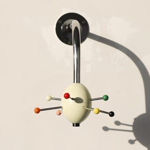 May include: A wall-mounted coat rack featuring a central cream-colored oval hub with multiple radiating metal arms. Each arm is tipped with a small sphere in various colors, including red, yellow, green, black, and pink, attached to a chrome stem.